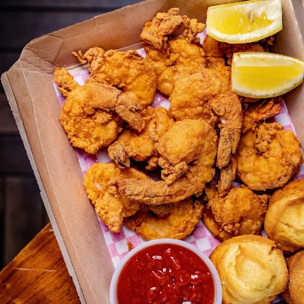 Crispy fried shrimp and hushpuppies with lemon wedges and a side of red sauce in a cardboard box.