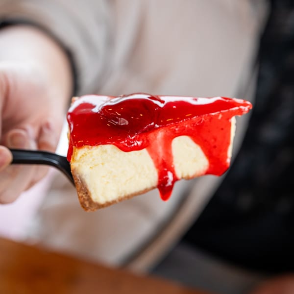 A hand holding a slice of cheesecake with a generous topping of red strawberry sauce.