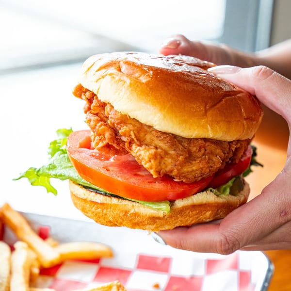 A hand holds a fried chicken sandwich with lettuce and tomato, next to fries in a red checkered basket.