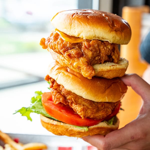 A hand holds a stacked fried chicken sandwich with lettuce, tomato, and cheese on a brioche bun.