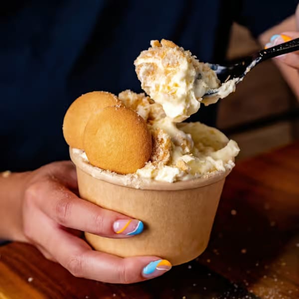 A person with colorful nails holds a cup of banana pudding with vanilla wafers, taking a spoonful.